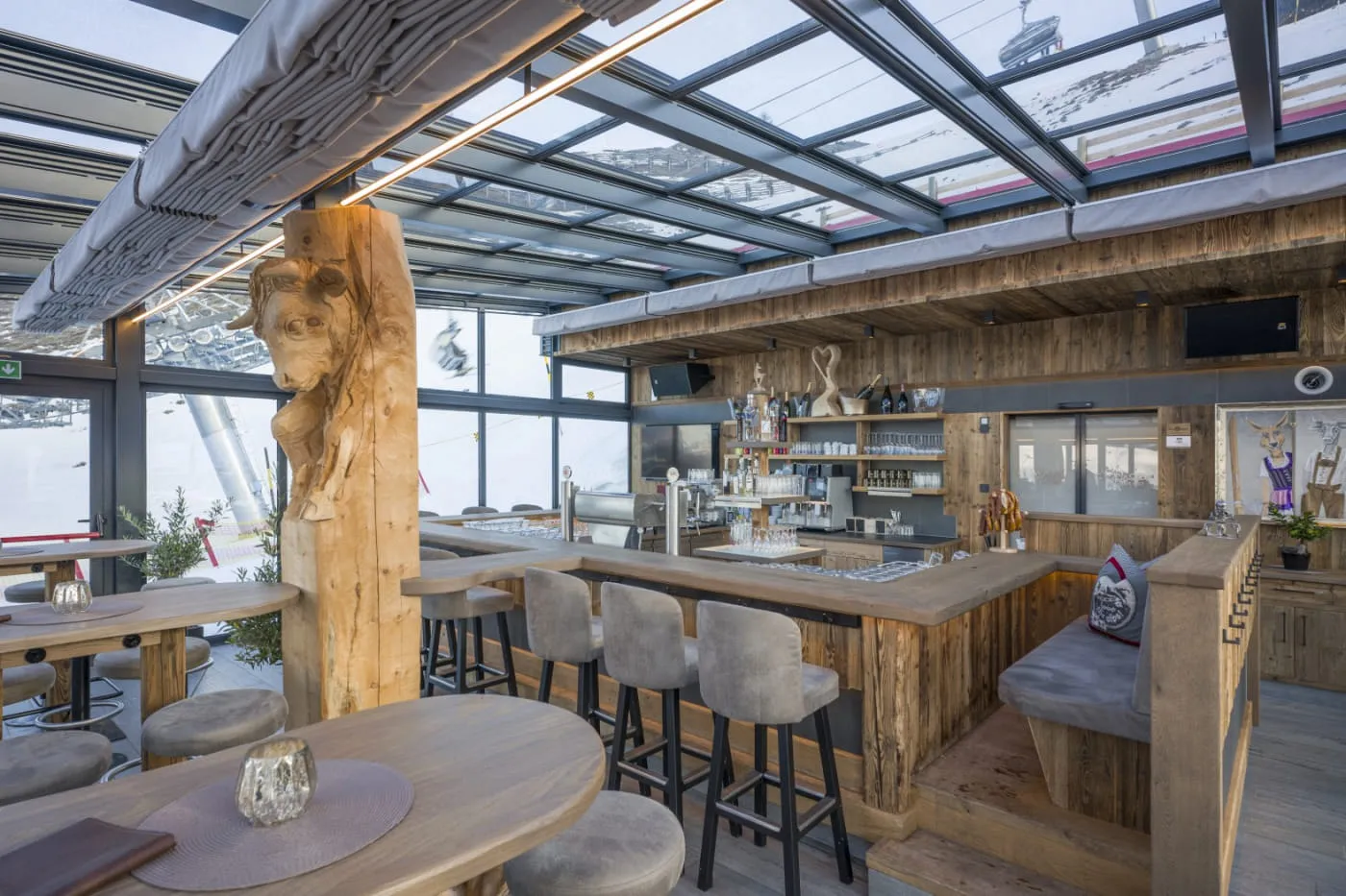 Wintergarten der OchsAlm mit Glasdach, Altholz-Bar und geschnitztem Ochsenkopf, dahinter die Skipiste
